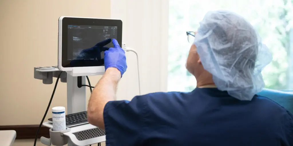 He is using his gloved finger to operate the touchscreen interface of a tablet-style ultrasound monitor